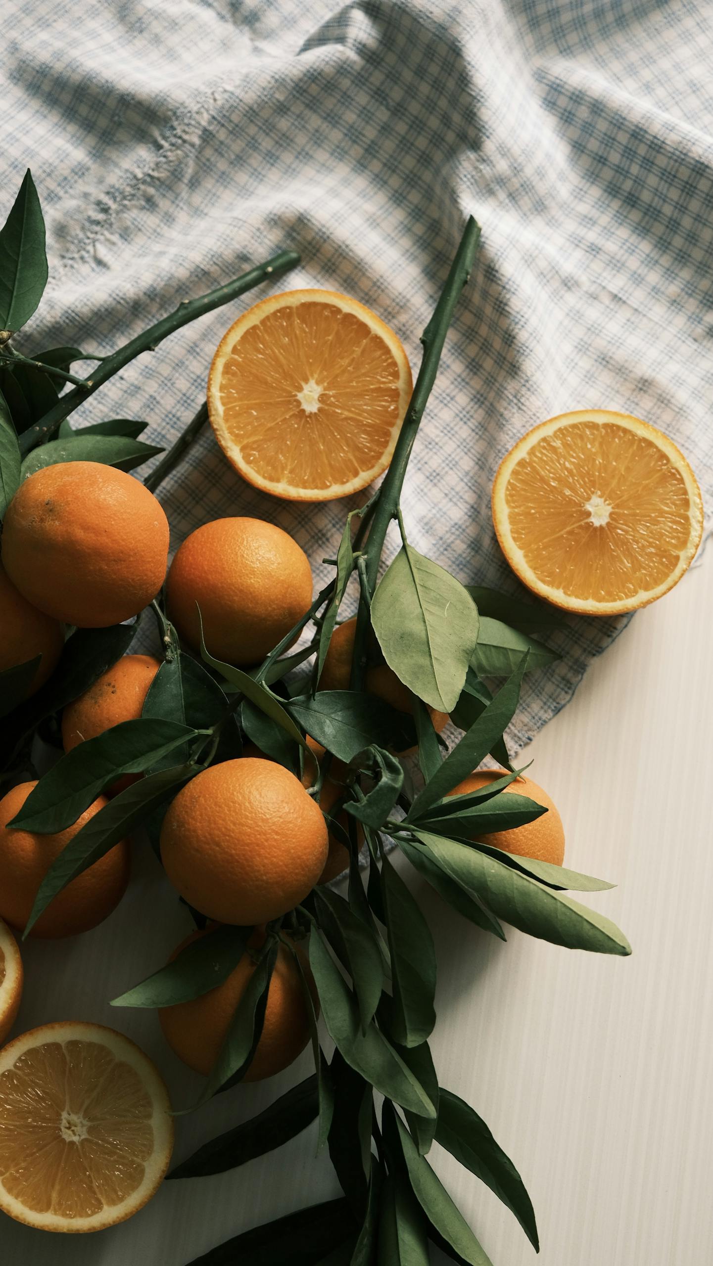 Fresh oranges with leaves on a checkered cloth, casting soft shadows.